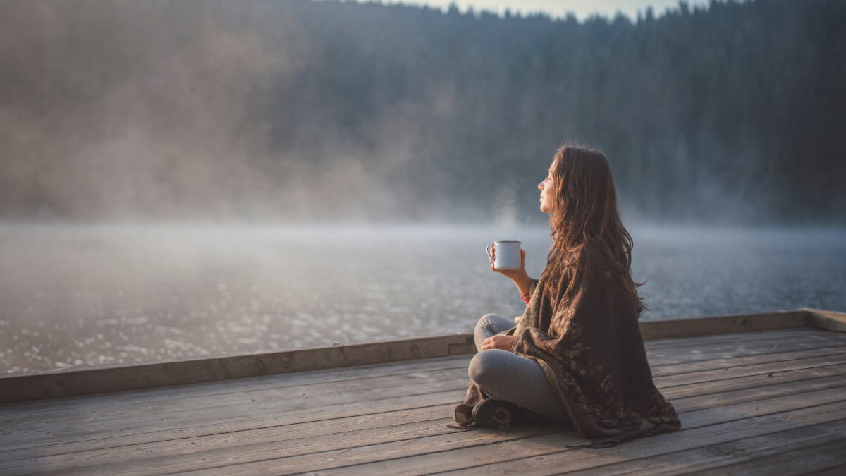 Woman sitting quietly on a dock on a foggy morning, holding a cup of coffee, reflecting peacefully — symbolizing gentle healing and intention-setting after narcissistic abuse.